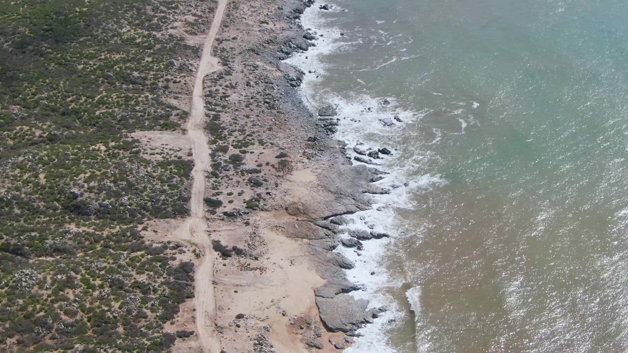 Aerial view at Buzzards Beach East Cape BCS Mexico with the dirt track centered near scrubland and waves breaking around dark shoreline rocks, with no visible people.