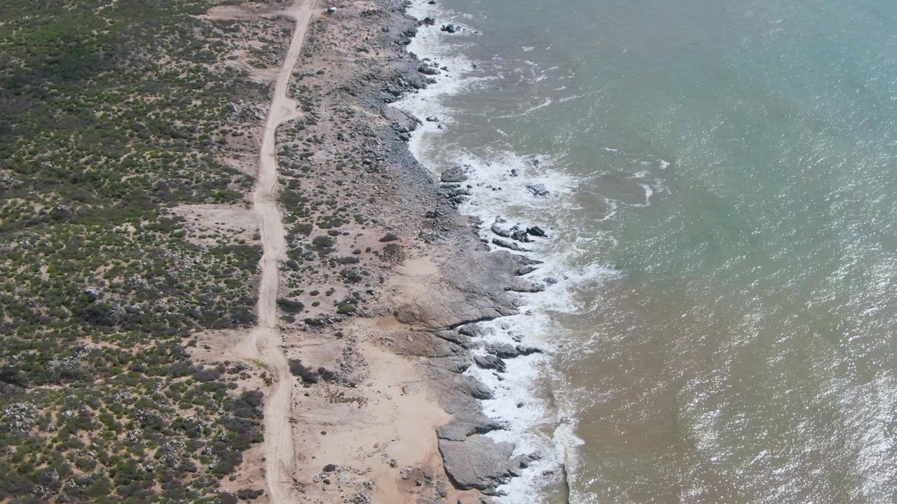 Aerial frame over Buzzards Beach East Cape BCS Mexico showing tan shallows, white surf, scattered rocks, sandy ground, a dirt road, and no visible people.
