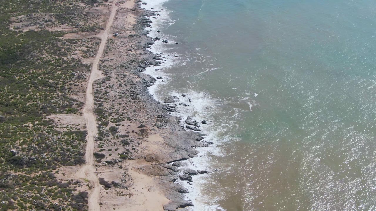 Aerial frame from Buzzards Beach East Cape BCS Mexico showing a dirt road along scrubland, rocky shoreline, white surf, and blue-green water with no visible people.