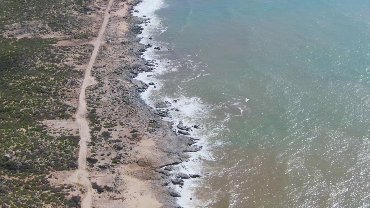At 0:01, the Buzzards Beach East Cape BCS Mexico shoreline remains angled through frame, with the dirt road on land, surf breaking over dark rocks, and no visible people.