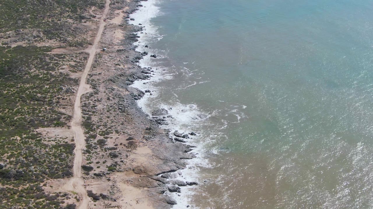 At 0:02, the aerial view shows more open water to the right of the Buzzards Beach East Cape BCS Mexico shoreline, with a pale dirt road and no visible people.