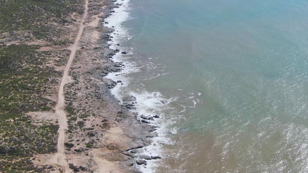 Rocky shoreline and turquoise water beside a dirt road through sparse vegetation.