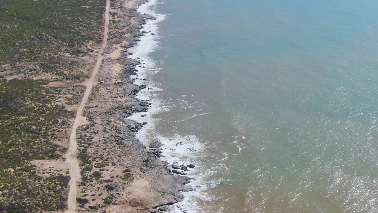 Aerial view of a dirt road beside a rocky coastline and pale surf.