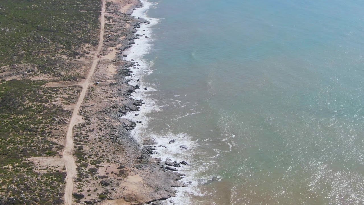 At one second, the aerial view holds the dirt road left of the rocky Buzzards Beach shoreline, with foamy waves spreading across gray-green water and no visible people.