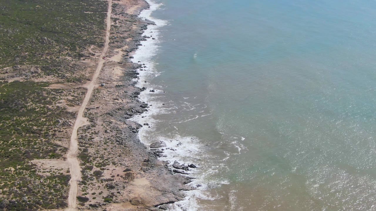 At three seconds, the aerial view shows more of the rocky shoreline and pale road through low vegetation at Buzzards Beach, with small surf lines in the water and no visible people.