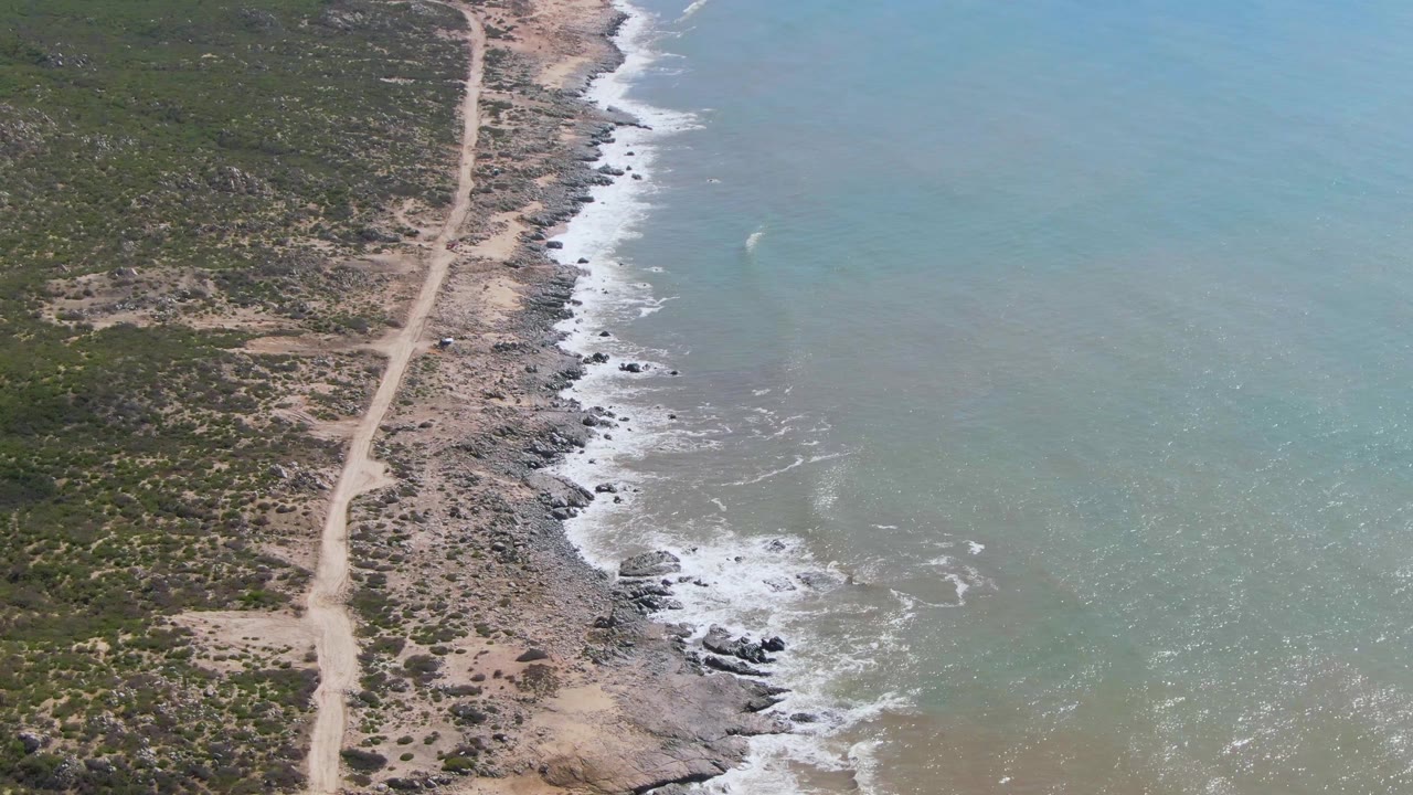 Aerial view of dirt road beside rocky coastline and turquoise ocean