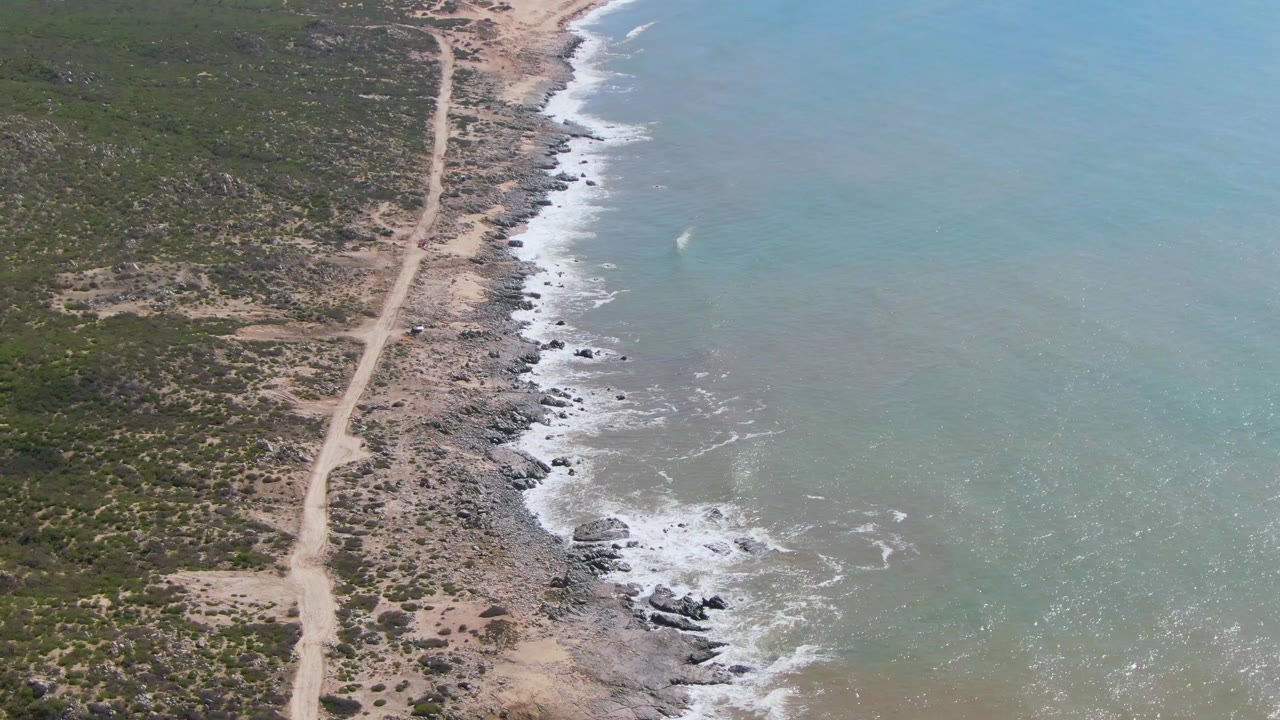 The second frame keeps the Buzzards Beach coastline centered as small waves break along dark rocks beside the pale dirt road and sparse green vegetation, with no visible people.