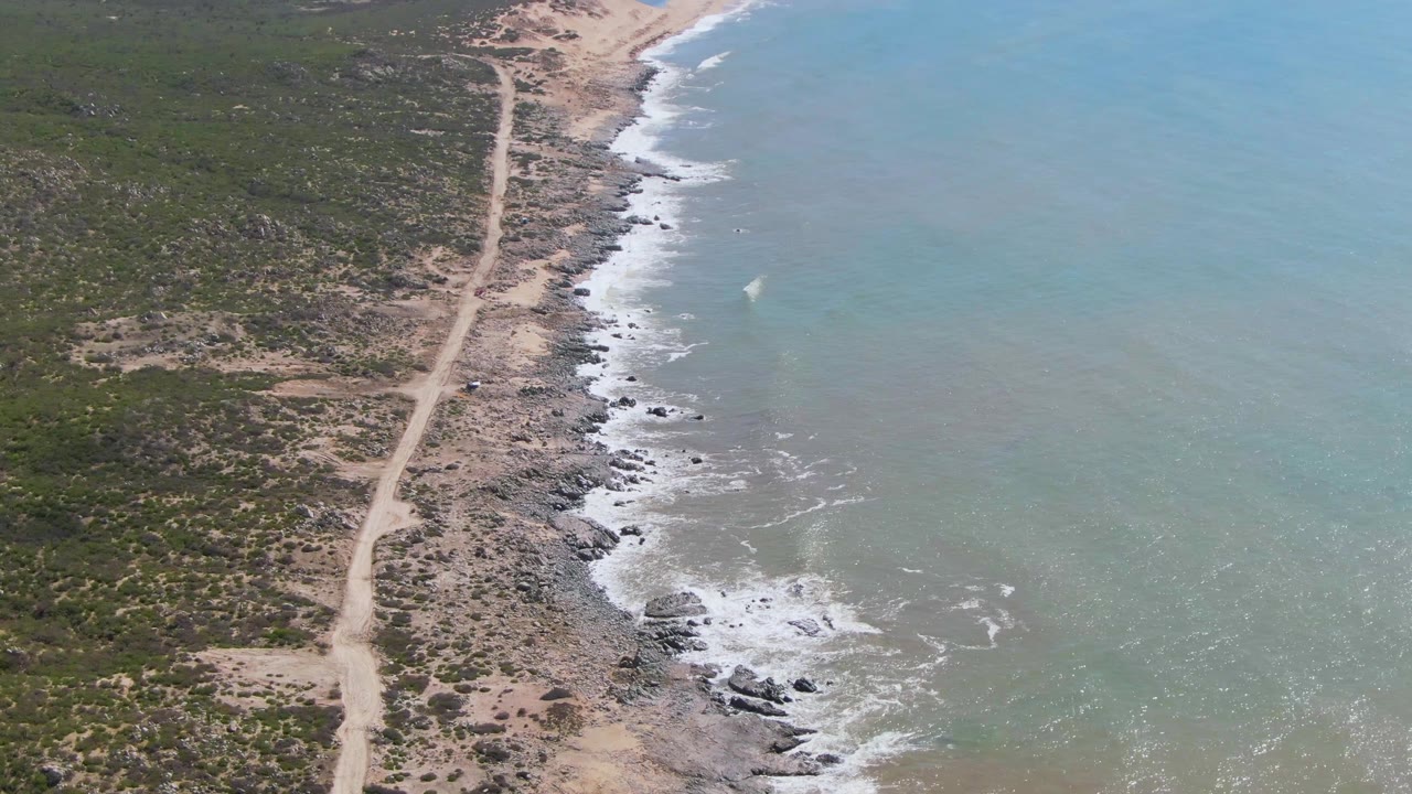At 0:02, the drone view shows more of the rocky Buzzards Beach shoreline extending upward through the frame, with pale road inland, blue-green ocean offshore, foam patches, and no visible people.