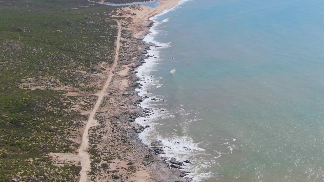 The 0:03 frame shows the dirt road bending slightly through green-brown coastal scrub while white water breaks over rocks along Buzzards Beach East Cape BCS Mexico, with no visible people.