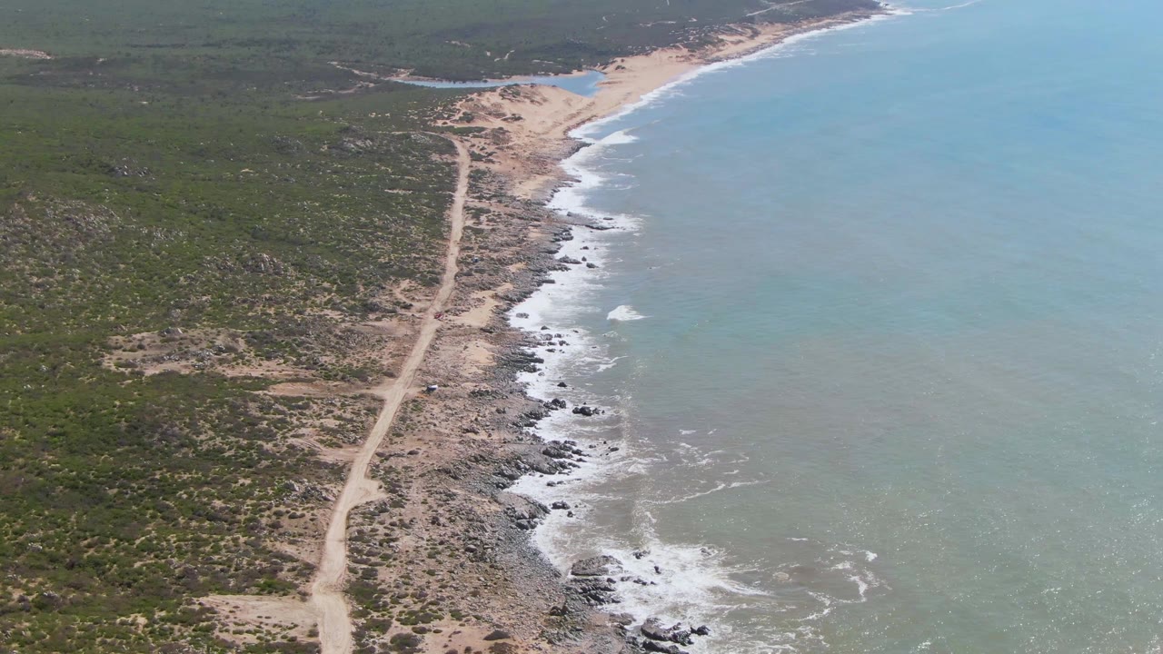 Aerial view over Buzzards Beach East Cape BCS Mexico showing a pale dirt road beside a rocky sandy shoreline, turquoise ocean, distant green hills, and no visible people.