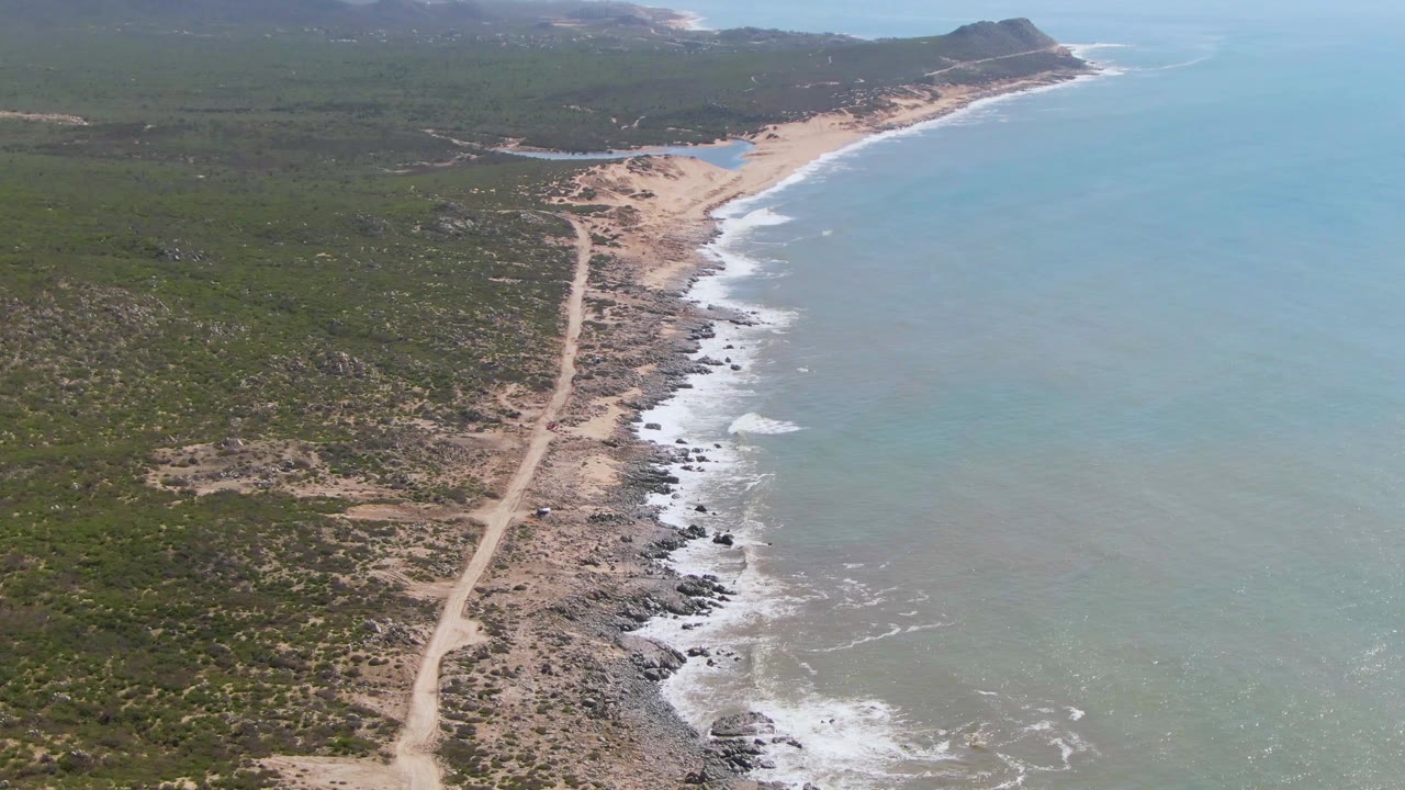 A wide aerial frame from Buzzards Beach East Cape BCS Mexico shows the road leading toward a sandy bend, surf whitening the rocky shore, open ocean on the right, low hills ahead, and no visible people.