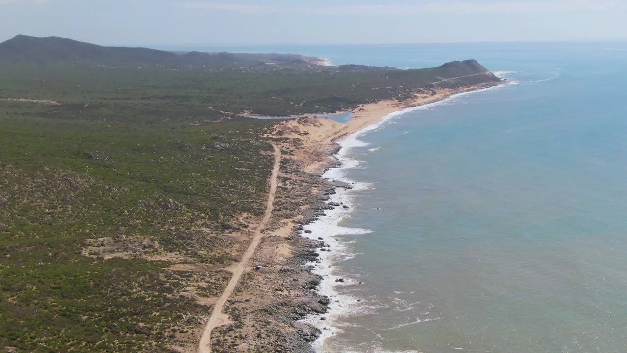 Aerial view of a sandy coastline with a dirt road and ocean surf.
