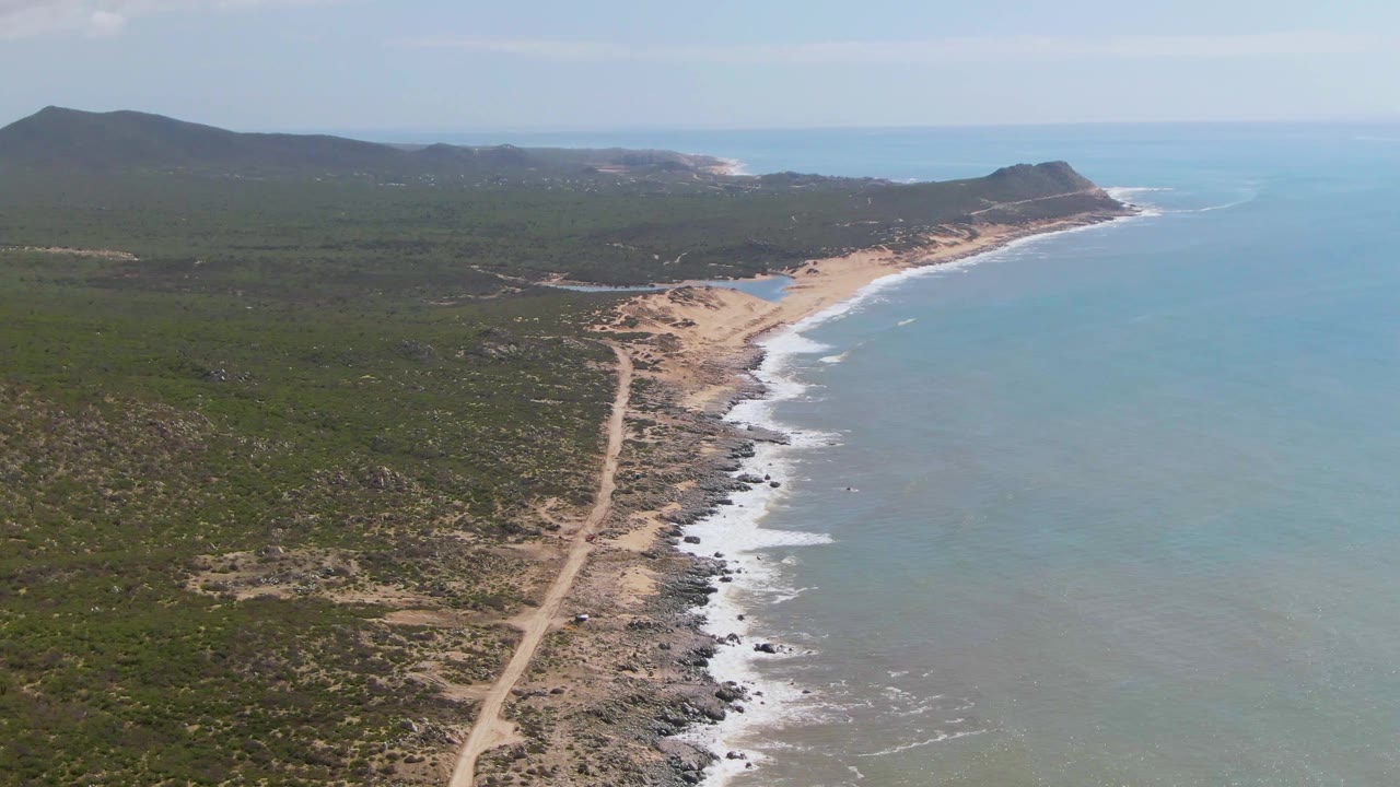 High oblique frame of Buzzards Beach East Cape BCS Mexico with scrub-covered land on the left, a sandy beach and surf line through the center, blue ocean on the right, and no visible people.
