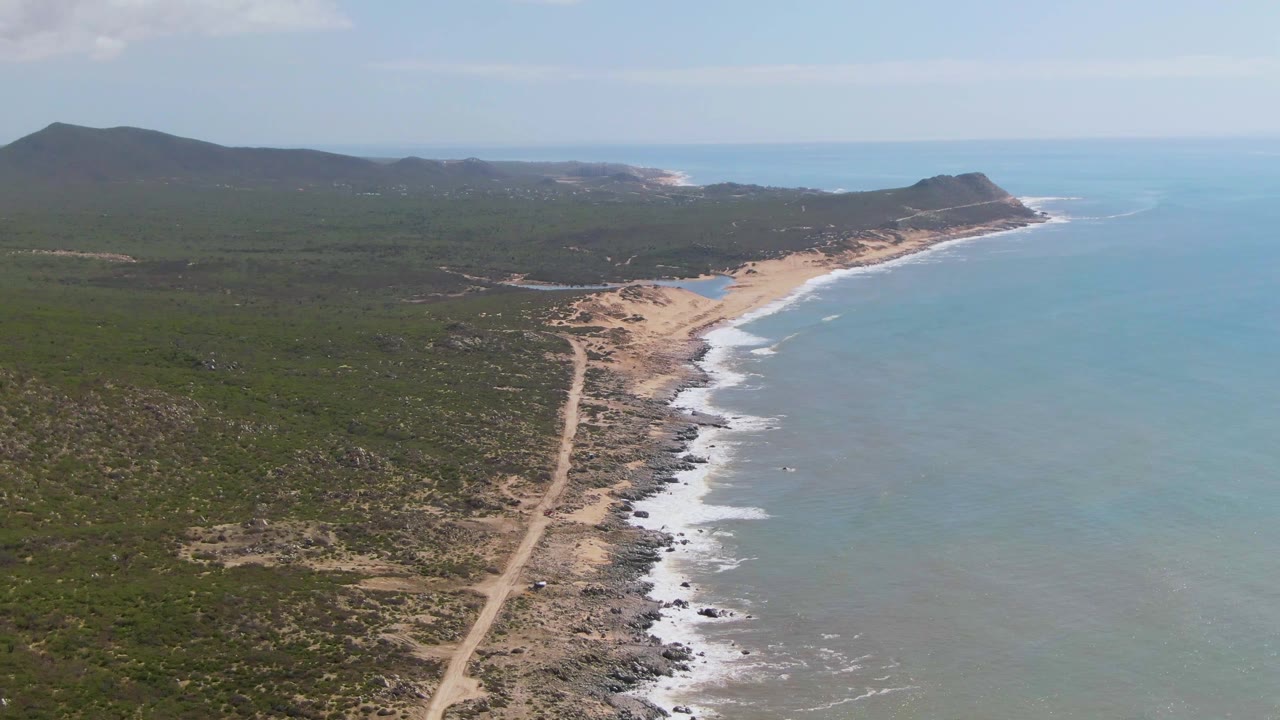 Aerial frame over Buzzards Beach East Cape BCS Mexico showing the dirt road meeting a sandy bend in the shoreline, with small white waves, green hills, and no visible people.