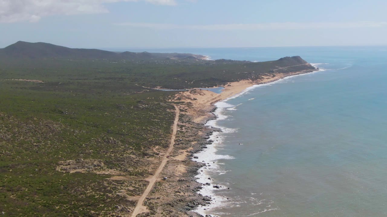 Wide aerial view of Buzzards Beach East Cape BCS Mexico with more open scrubland visible in the foreground, a dirt road leading to the sandy beach, ocean surf, and no visible people.