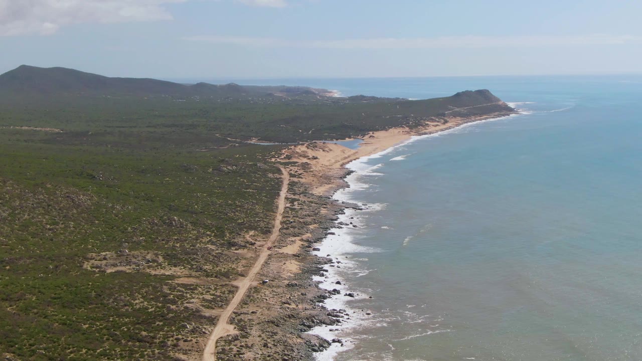 Aerial footage over Buzzards Beach East Cape BCS Mexico shows the road running closer to the center of frame, with surf bands curling along the shore and no visible people.