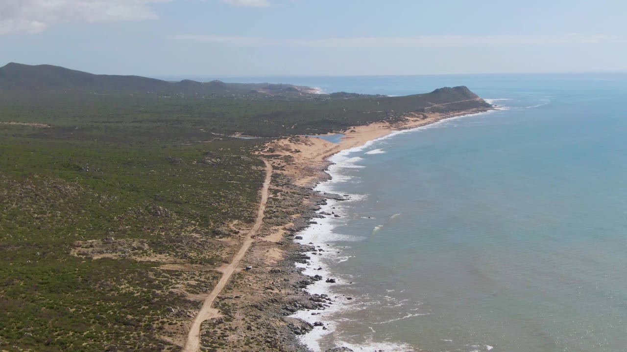 A later aerial frame over Buzzards Beach East Cape BCS Mexico shows the dirt track bending through scrubland toward a rocky beach, with pale blue water and no visible people.