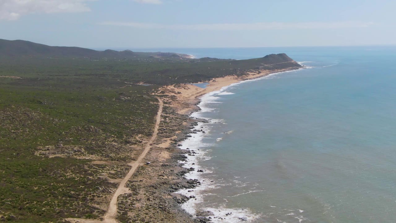 Aerial view of Buzzards Beach East Cape BCS Mexico with a dirt road beside scrubland, a sandy and rocky shoreline, blue-green ocean, and no visible people.