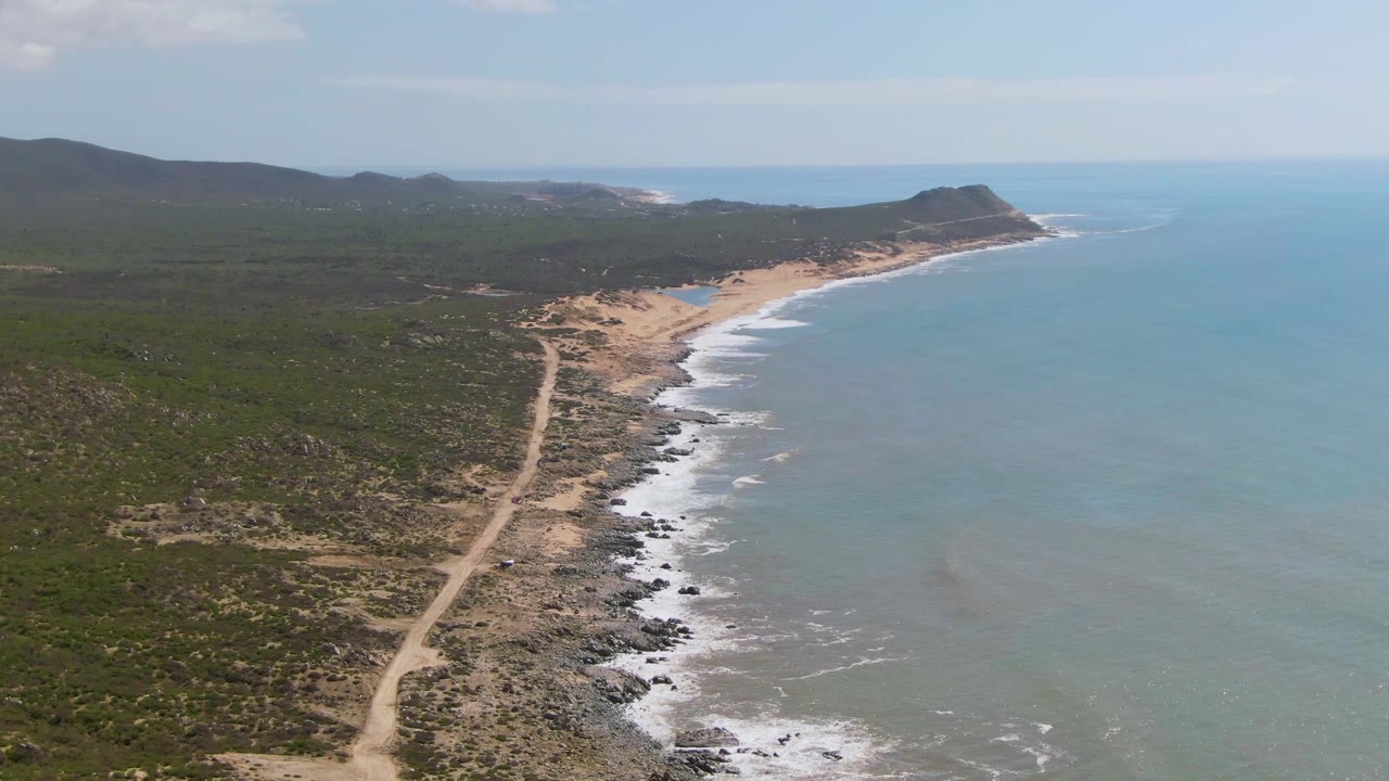 A second aerial frame over Buzzards Beach East Cape BCS Mexico shows the road running toward the beach while white wave lines advance along the shore, with no visible people.