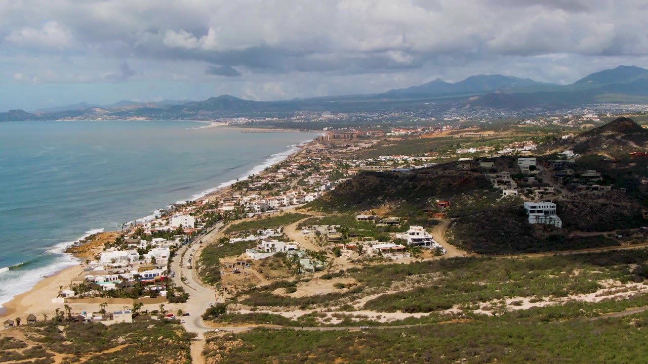 Aerial view over Buzzards Beach East Cape BCS Mexico with turquoise ocean at left, sandy beach and coastal road below, white houses on scrubby hills, distant mountains, cloudy sky, and no visible people.
