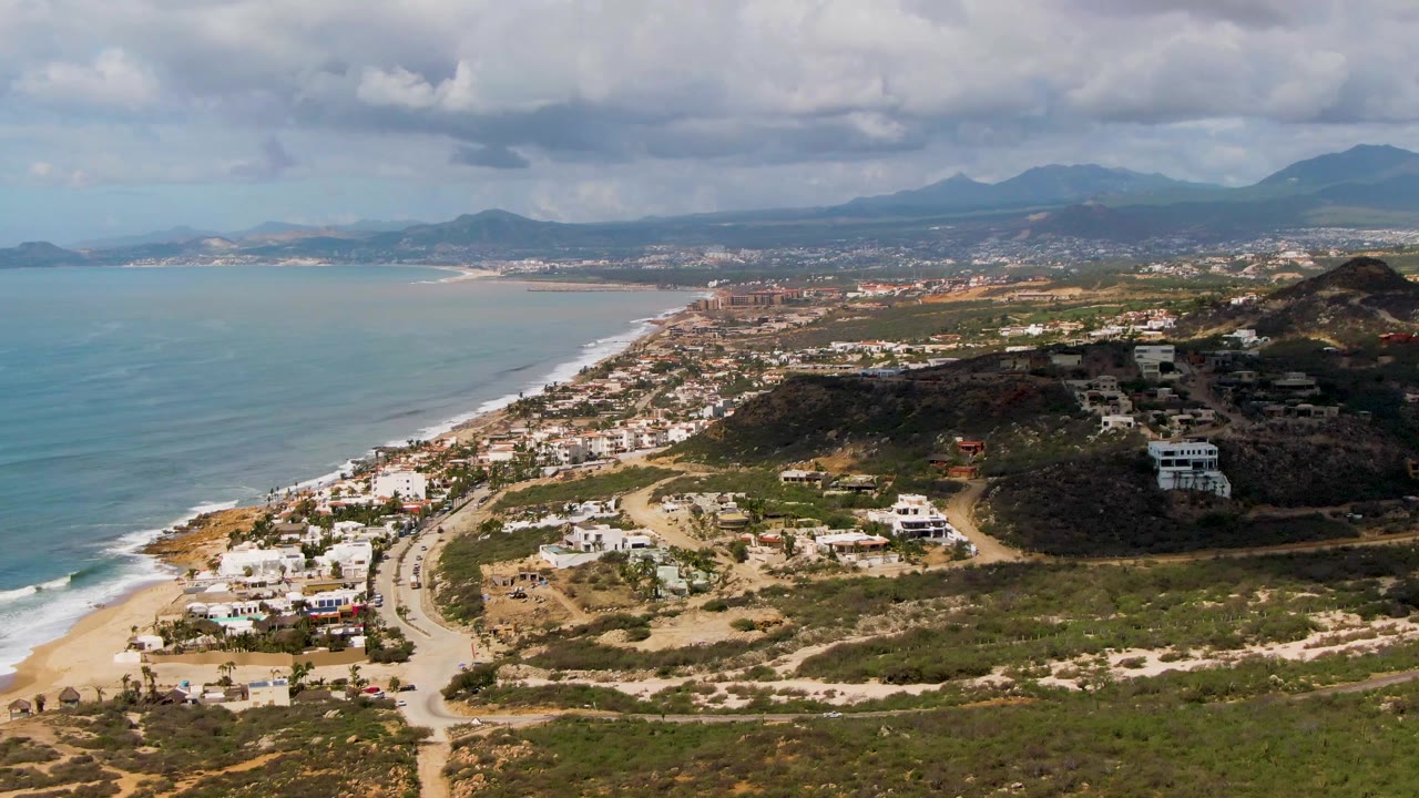 Coastal aerial with ocean, beach road, hillside houses, green scrub, and mountains.