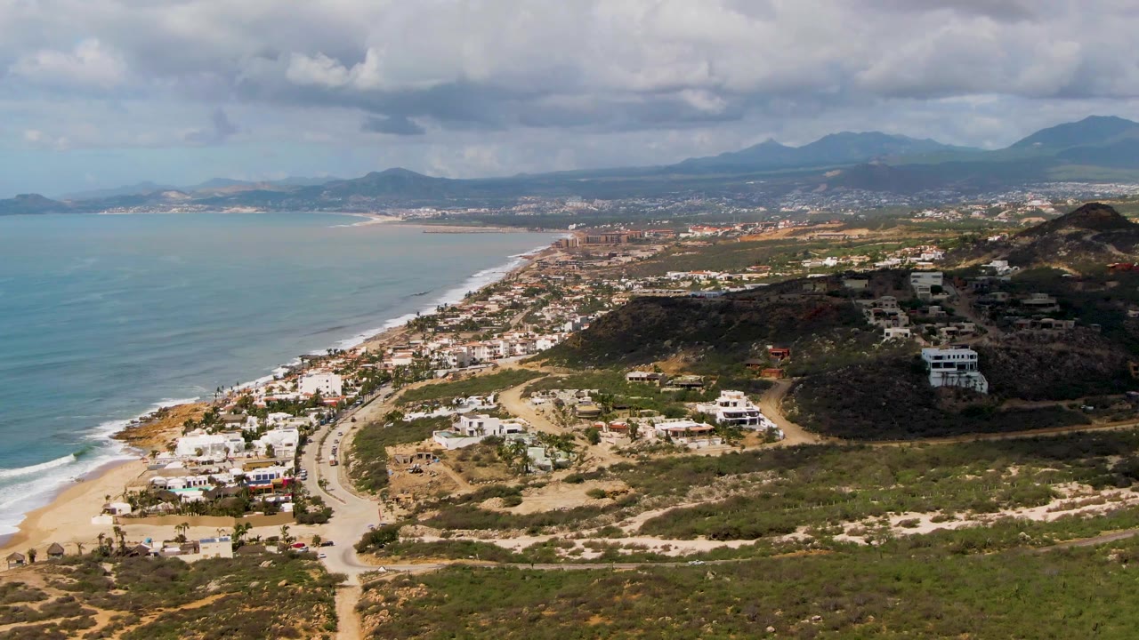 Wide drone view over Buzzards Beach East Cape BCS Mexico with pale surf along the beach, a road near the coast, white homes on brown-green terrain, mountains in the distance, heavy clouds, and no visible people.