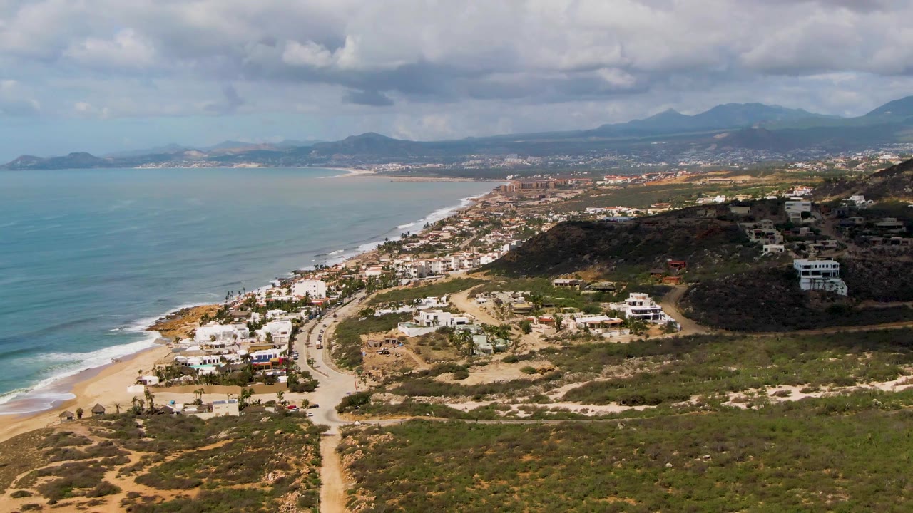 Opening aerial frame over Buzzards Beach East Cape BCS Mexico with ocean surf at left, sandy beach and road below, hillside buildings at right, distant mountains, heavy clouds, and no visible people.