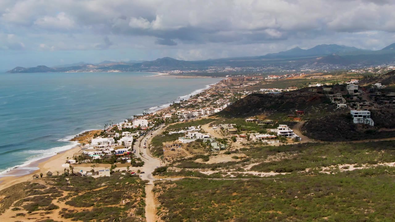 Aerial frame from Buzzards Beach East Cape BCS Mexico showing surf bands along the left shore, a sandy access road through brush, hillside homes on the right, mountains in the distance, cloud shadows, and no visible people.