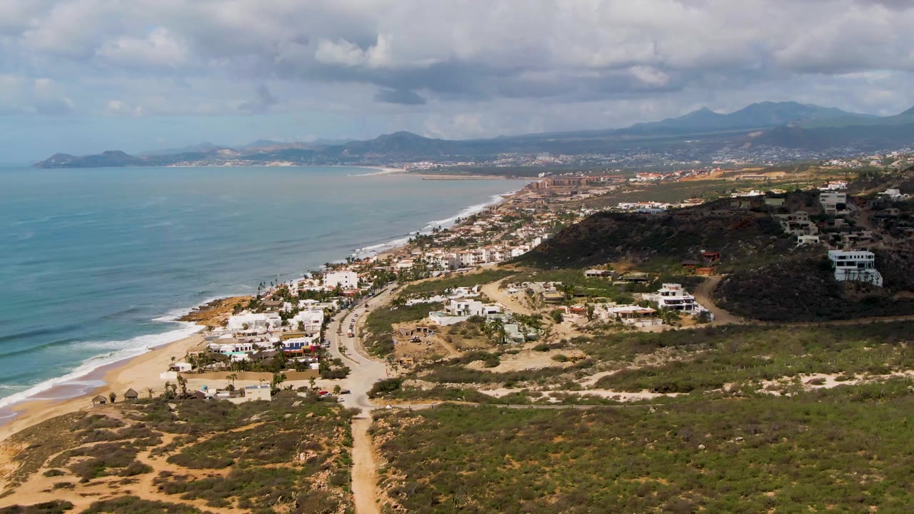 Mid-clip aerial view at Buzzards Beach East Cape BCS Mexico with ocean waves rolling toward the beach, scattered white buildings above scrubland, a road cutting through the foreground, distant mountains, and no visible people.