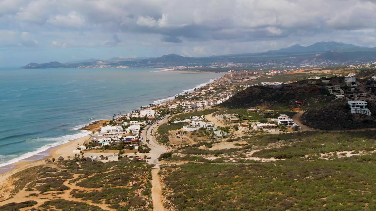 Coastal aerial scene with ocean on the left, beach road below, homes on hills, and clouded mountains.