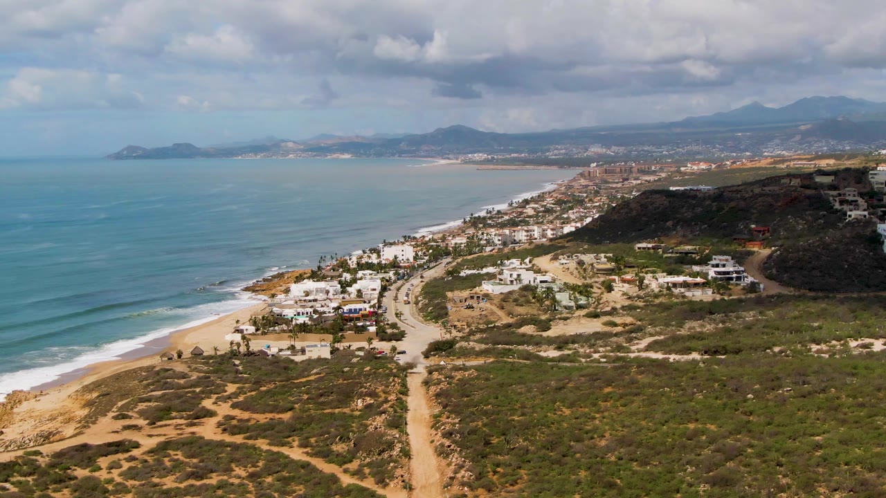 Aerial view over Buzzards Beach East Cape BCS Mexico, with turquoise ocean at left, a sandy beach, scrubland paths, scattered homes, hills, distant mountains, heavy clouds, and no visible people.