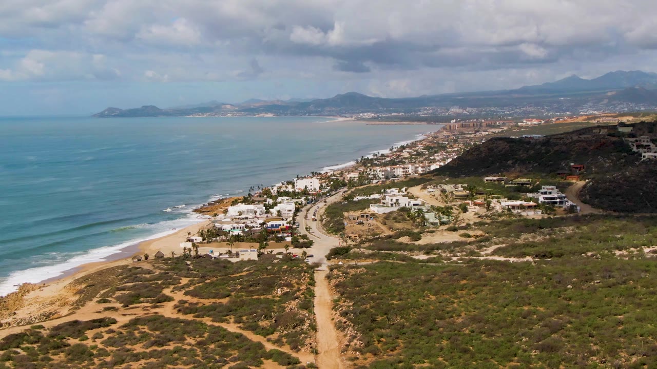 High aerial coastline view with ocean, sandy beach, dirt road, white buildings, hills, and clouds.