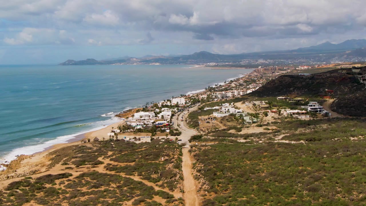 Aerial frame from Buzzards Beach East Cape BCS Mexico with the surf line lower left, beach houses near the center, scrubland and a dirt track in front, distant town and mountains beyond, and no visible people.