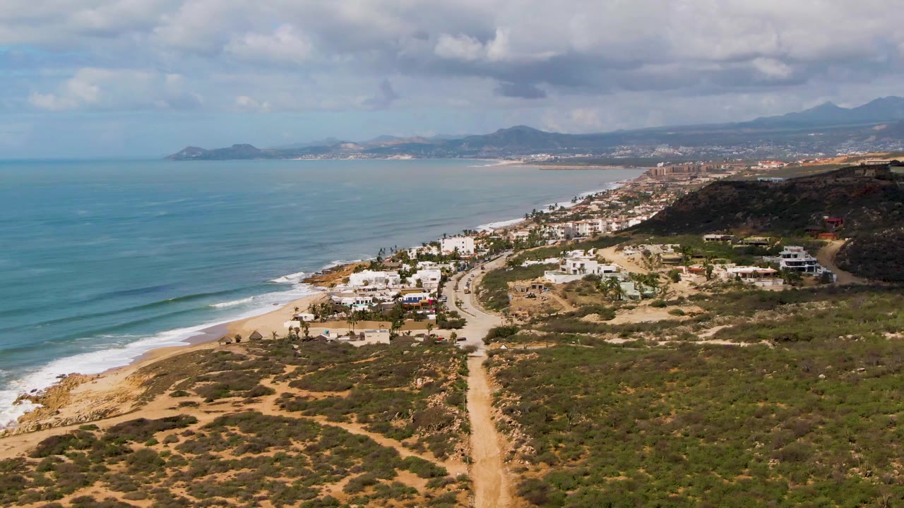 Aerial coastal landscape with blue ocean, pale sand, low waves, scrub vegetation, houses, and mountains.