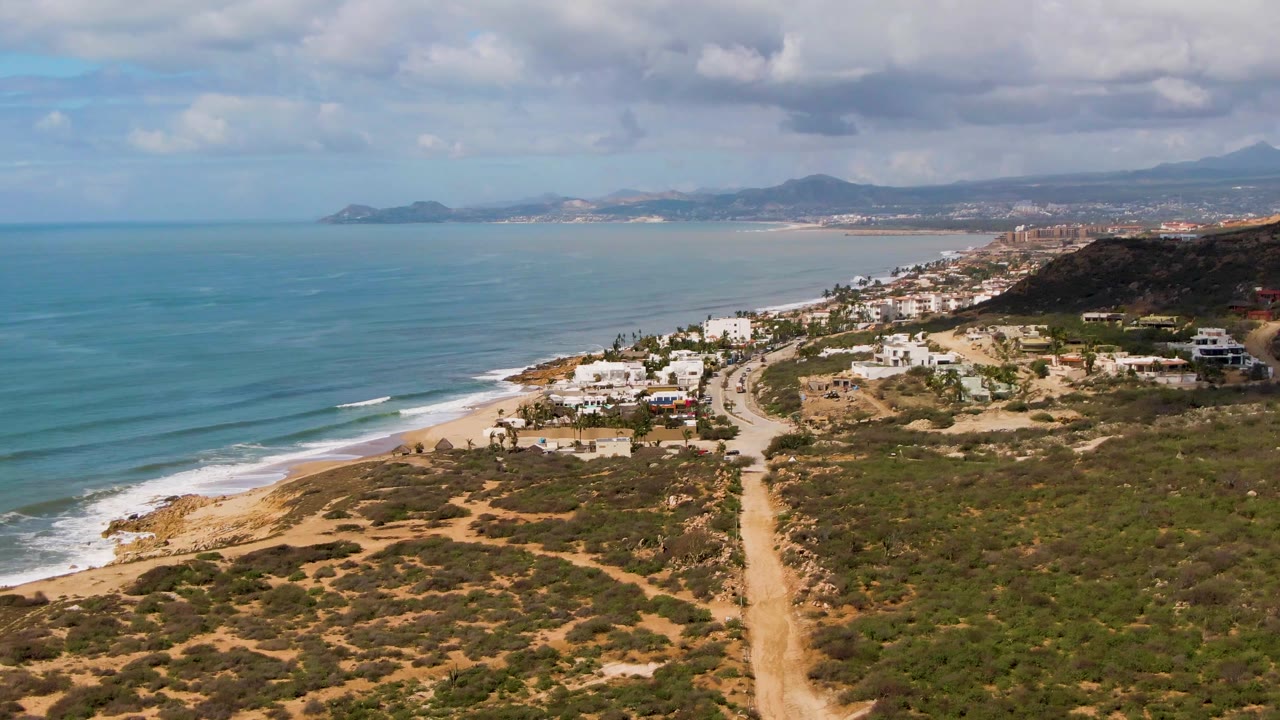 Aerial frame over Buzzards Beach East Cape in Mexico showing a tan dirt road through scrubland, white coastal houses, blue ocean, distant hills, clouds, and no visible people.