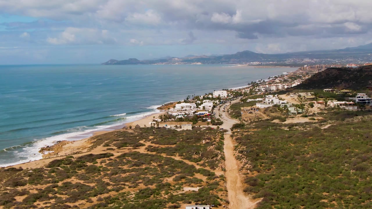 Aerial view over Buzzards Beach East Cape in Mexico with the road centered more clearly, surf breaking along the beach, scattered white buildings, distant hills, cloud cover, and no visible people.
