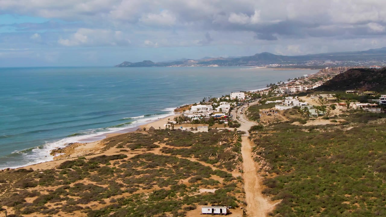 Aerial frame from Buzzards Beach East Cape in Mexico showing scrub-covered terrain, a straight dirt road near frame center, white houses near the beach, rolling ocean lines, distant hills, and no visible people.