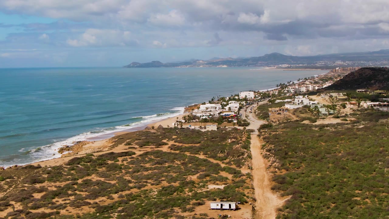 Wide aerial coastline with dirt road, surf, homes, and distant mountains.