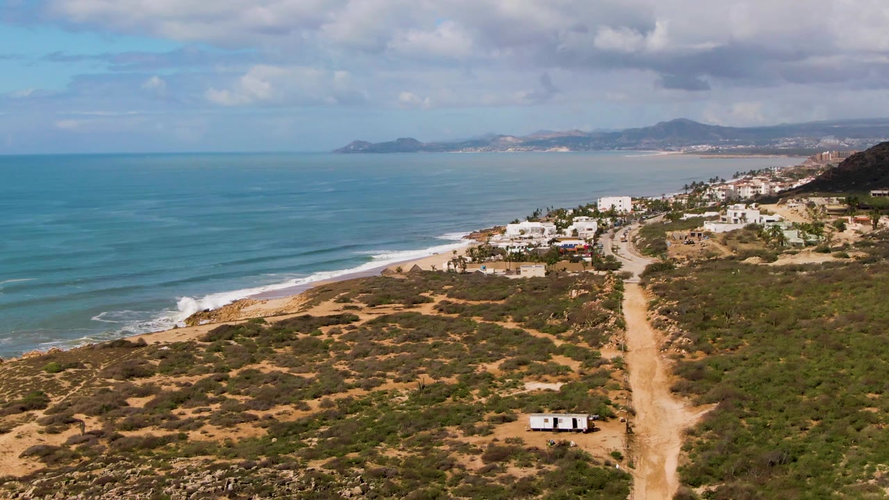 Aerial frame at Buzzards Beach East Cape BCS Mexico showing a dirt road through scrubland, white coastal buildings, blue ocean and surf to the left, distant hills, and no visible people.