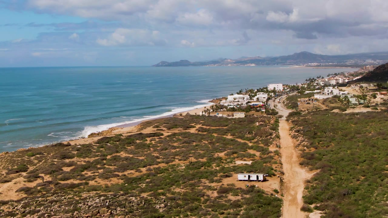 Coastal aerial with blue ocean, beach, dirt road, and hillside buildings.