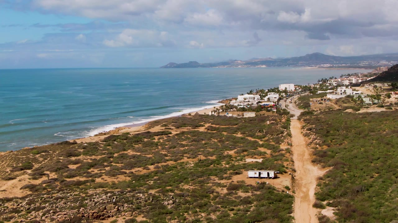 Aerial frame at Buzzards Beach East Cape BCS Mexico showing more of the road corridor on the right, a small white object near the lower road area, blue water on the left, and no visible people.