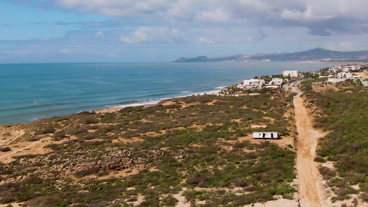 Aerial frame at Buzzards Beach East Cape BCS Mexico showing a sandy road through dry scrub, a small white trailer near the road, blue ocean at left, white buildings on the ridge, and no visible people.