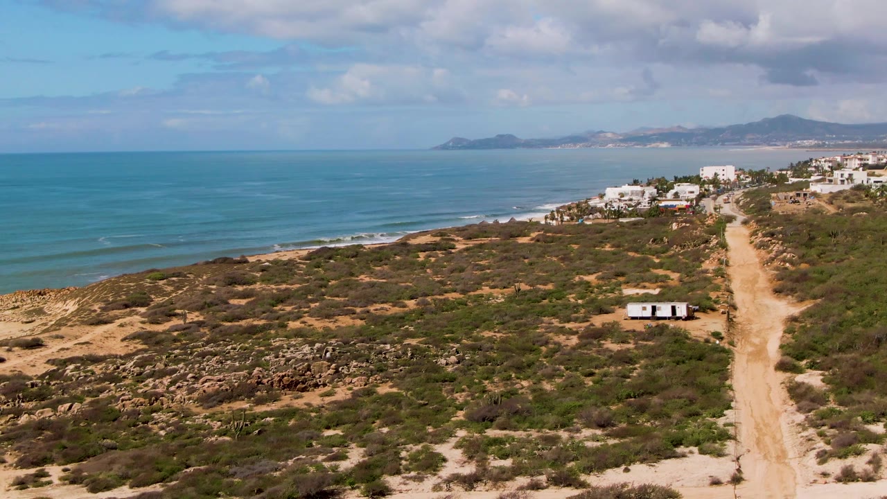 Aerial frame at Buzzards Beach East Cape BCS Mexico with the dirt road slightly more centered, the small trailer beside scrub vegetation, calm ocean waves along the shore, and no visible people.