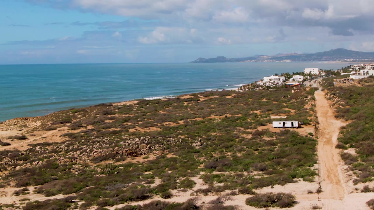 Aerial frame at Buzzards Beach East Cape BCS Mexico showing the ocean occupying the left half, dry scrub and rock patches in the foreground, the sandy road at right, the small trailer below the buildings, and no visible people.