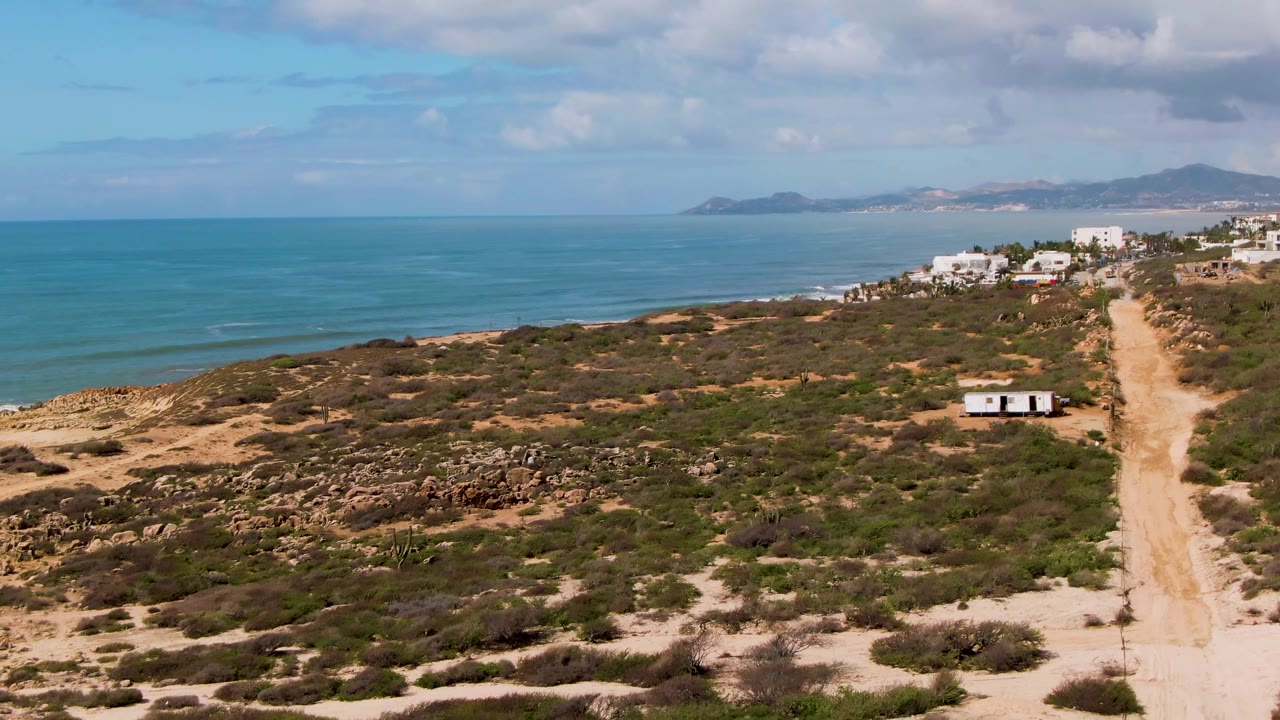 Coastal scrub and sandy road seen from above with ocean and white buildings beyond.