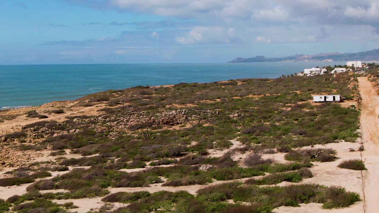 Opening frame of Buzzards Beach East Cape BCS Mexico with sandy scrubland in front, a straight dirt road on the right, small white buildings near the coast, blue ocean, distant land, and no visible people.