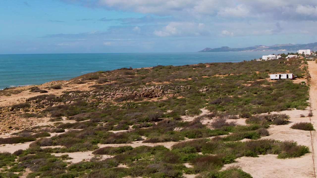 Second frame over Buzzards Beach East Cape BCS Mexico showing the dirt road tighter to the right edge, scrub-covered dunes across the center, ocean on the left, distant land on the horizon, and no visible people.