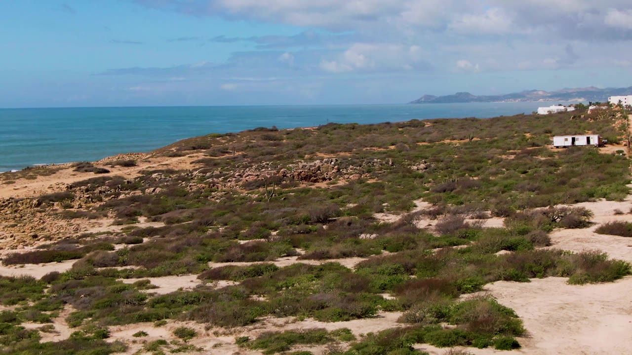 Third frame of Buzzards Beach East Cape BCS Mexico with more scrubland and sandy openings across the foreground, the road mostly at the far right, white buildings near the upper right, ocean behind them, and no visible people.