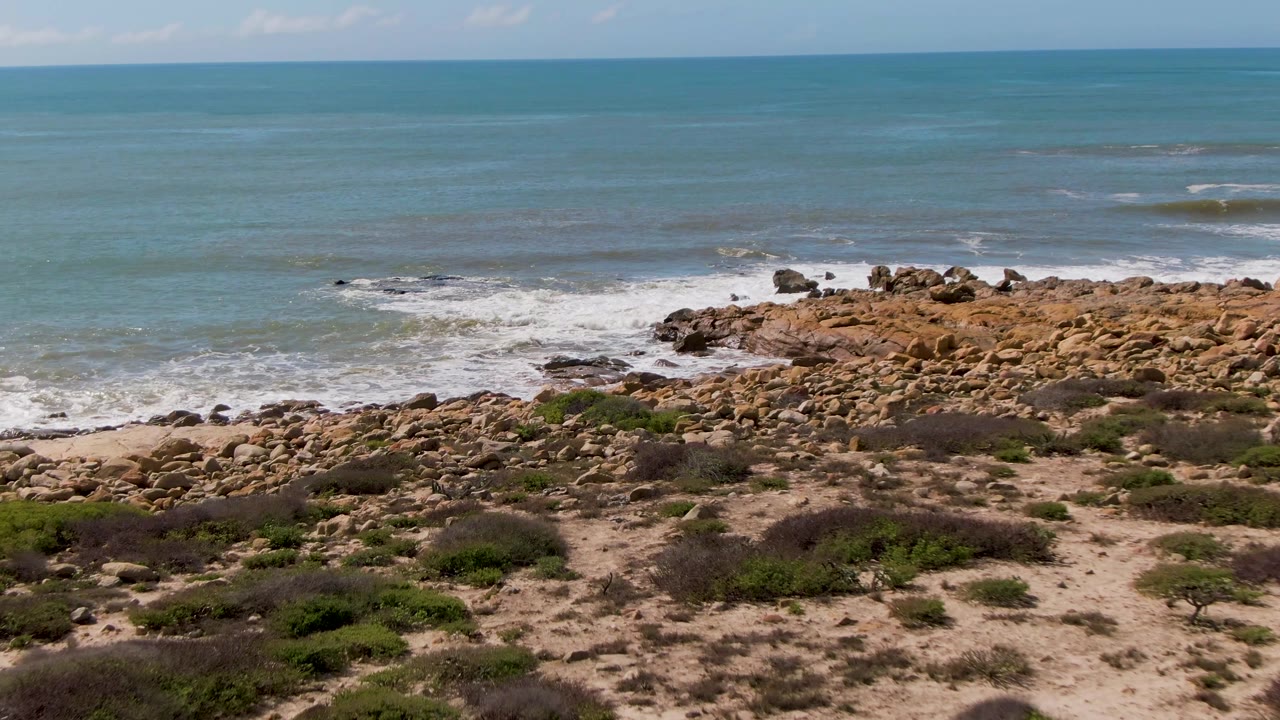 Opening frame at Buzzards Beach, East Cape BCS, Mexico, with low shrubs and sand in front of tan rocks, white surf, open ocean, and no visible people.