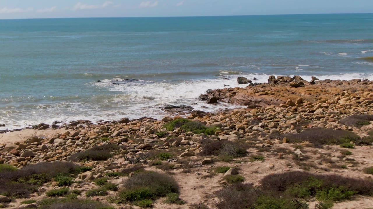 At 0:01, Buzzards Beach shoreline shows white water spreading around the rocks, with sandy ground and sparse green shrubs still visible in the lower frame and no visible people.